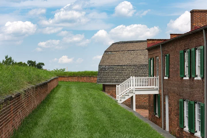 A brick barracks building with green shutters and a white wooden stair, below the grassy ramparts of the star fort