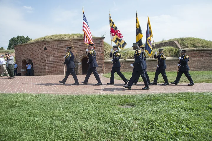 A color guard carrying U.S., Maryland and military flags marches across the brick plaza by the star fort