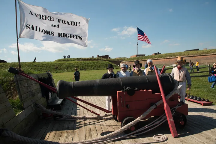 A black cannon on a red wooden carriage beside a white flag reading Free Trade and Sailors Rights, with the U.S. flag behind