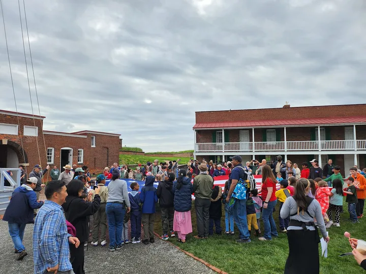 A crowd on the parade ground inside the star fort gathering around the flag during a flag change, brick barracks behind