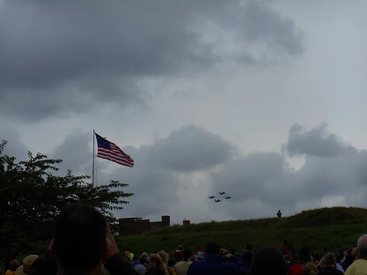 The U.S. flag flying over the ramparts of Fort McHenry under grey cloud, with a formation of jets passing overhead and a crowd below