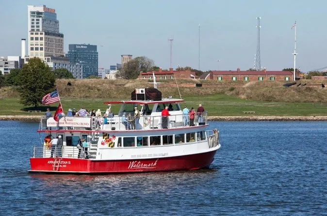A red and white Watermark cruise boat on the water with the grassy ramparts of Fort McHenry behind it