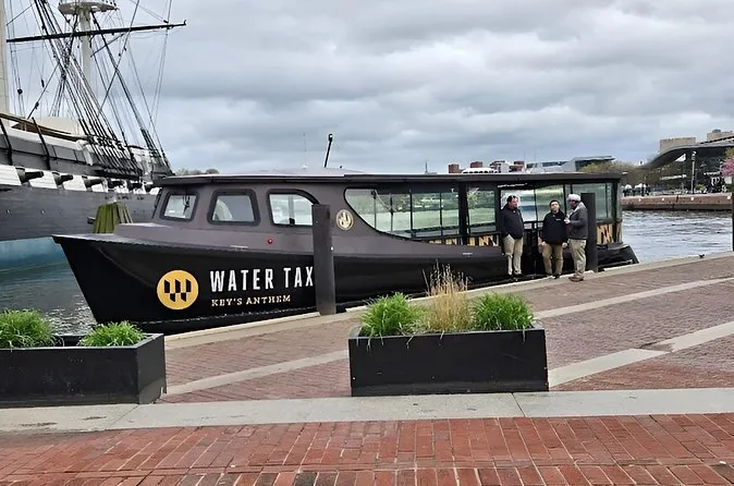 A black Baltimore Water Taxi boat moored at a brick promenade, with the masts of a tall ship behind